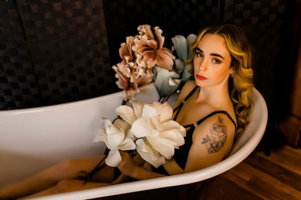 A woman immersed in a bathtub adorned with flowers in a Boudoir Gallery setting.