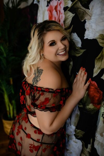 A woman in a red and black lace dress posing against a floral wall at a Boudoir Gallery.