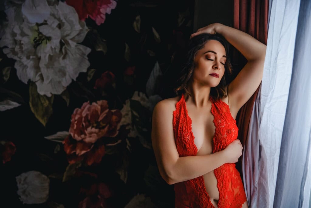 A woman in a red lingersuit leaning against a window.